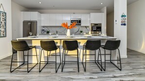 Una cocina moderna con armarios blancos, una isla central con taburetes de bar, electrodomésticos de acero inoxidable y decoración mural.