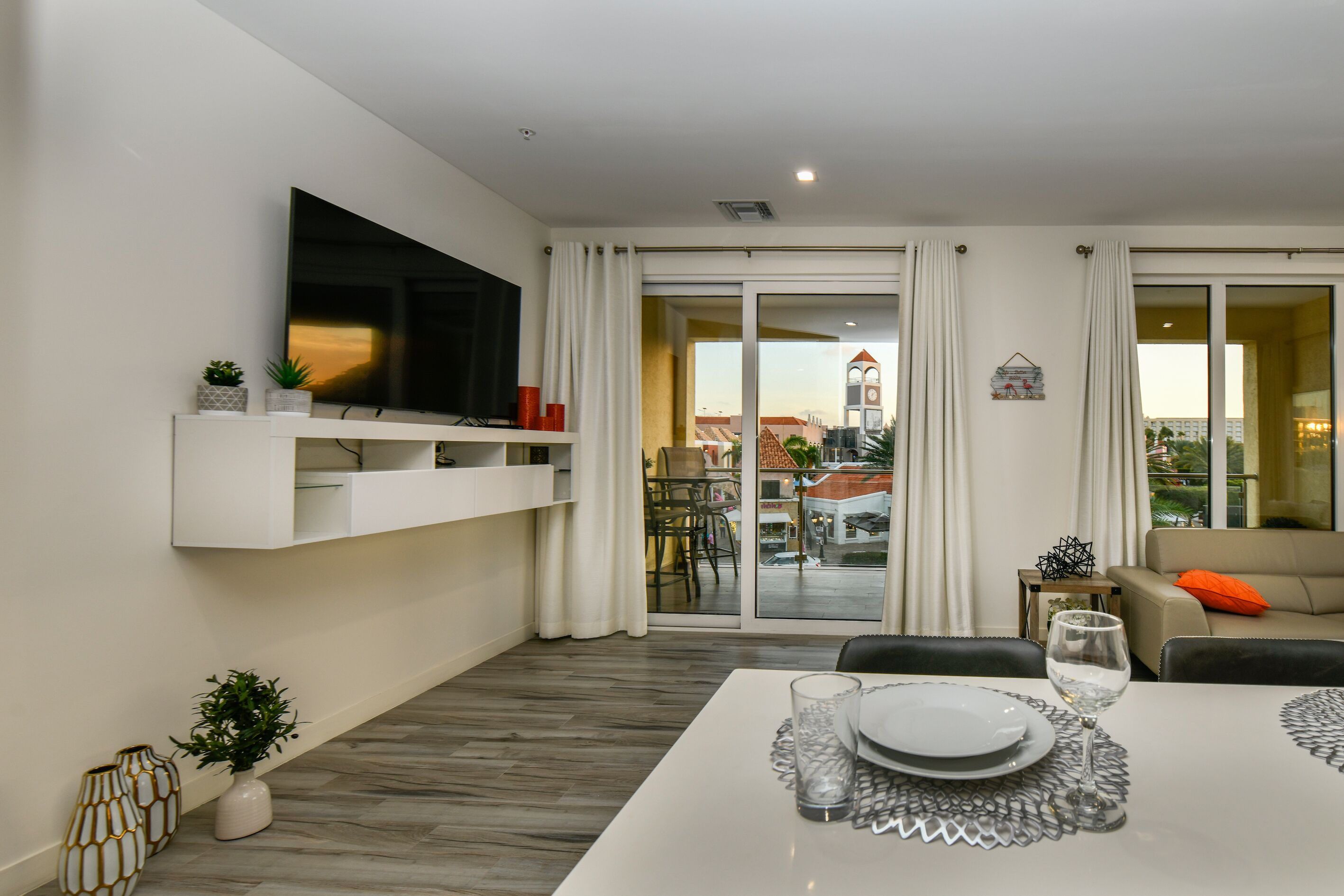 Un salon moderne doté d’un téléviseur à écran plat, d’une table à manger dressée avec des assiettes et des verres, et d’un canapé agrémenté d’un coussin orange.