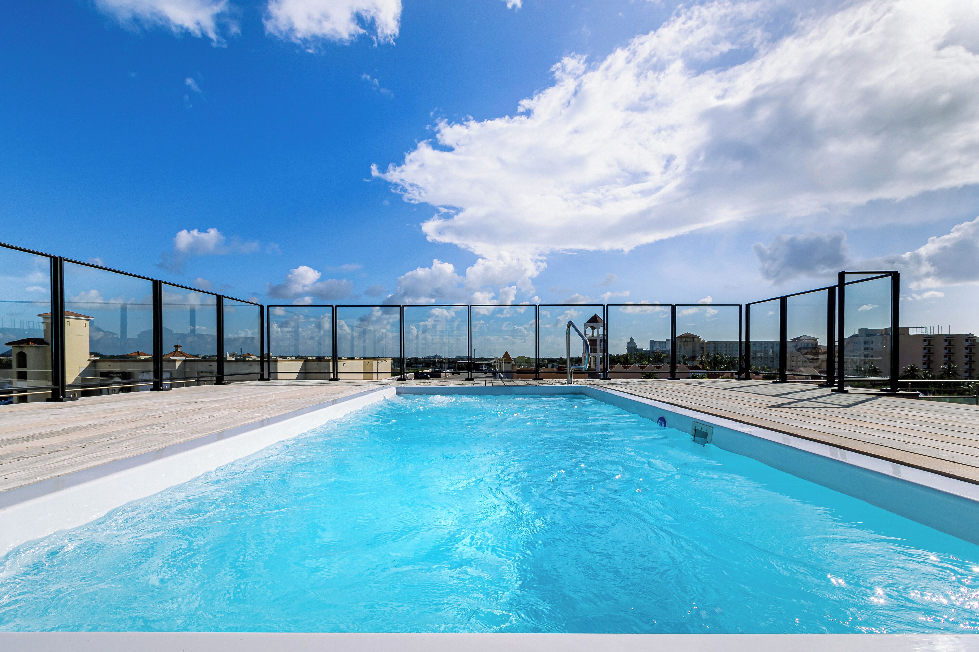 A rooftop swimming pool with clear blue water, surrounded by glass railings and a wooden deck.