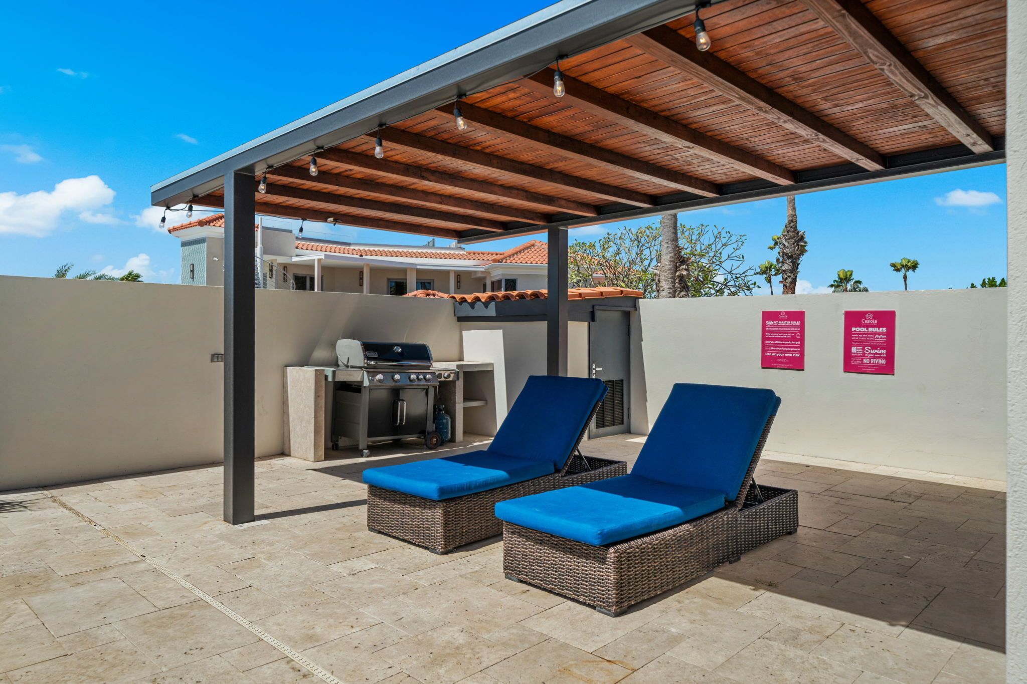 A covered outdoor area with two lounge chairs, a barbecue grill, and a wall with pool rules signs.