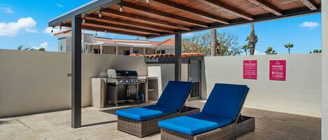 A covered outdoor area with two lounge chairs, a barbecue grill, and a wall with pool rules signs.