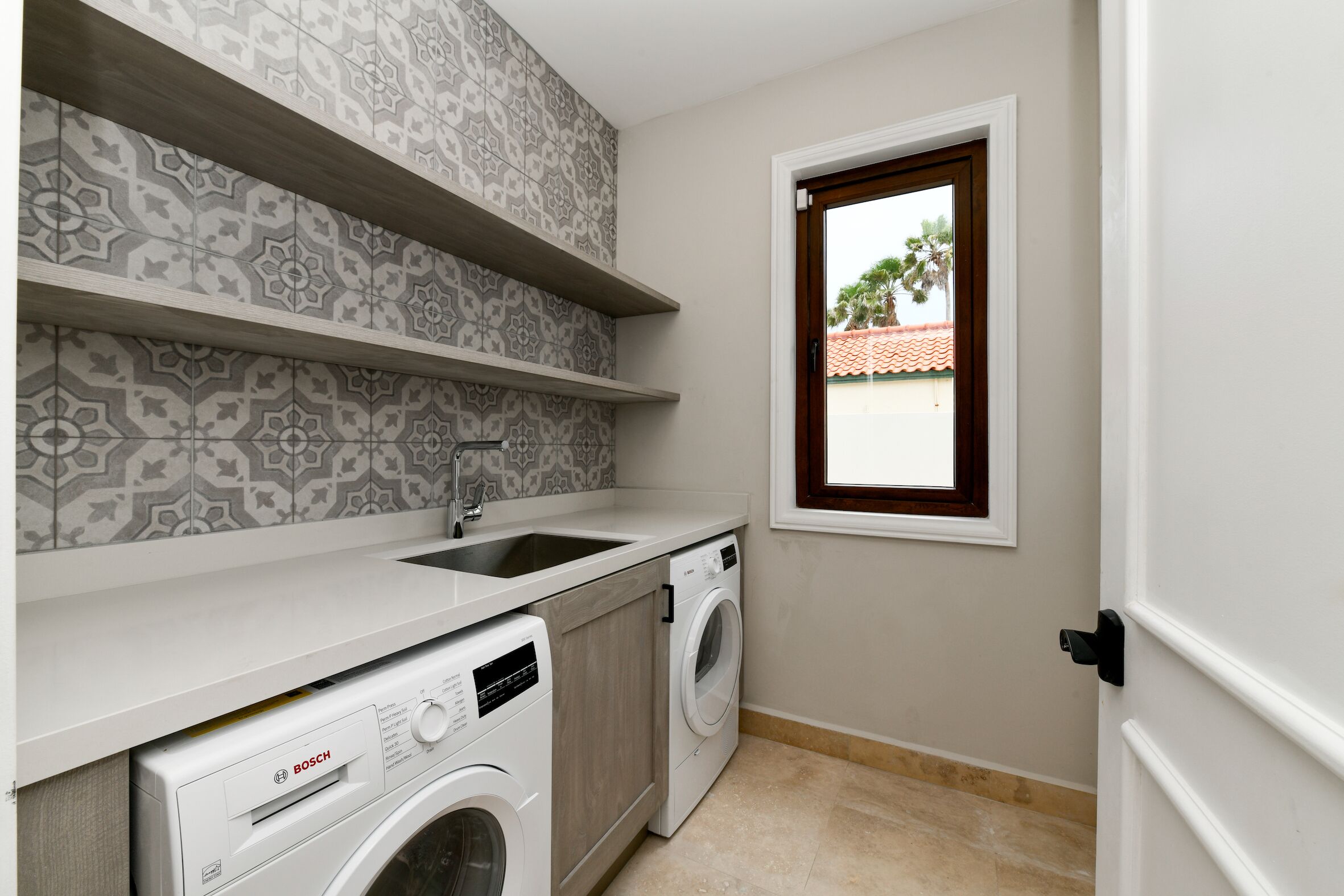 A compact laundry room with a Bosch washing machine, a sink, and patterned tile walls.
