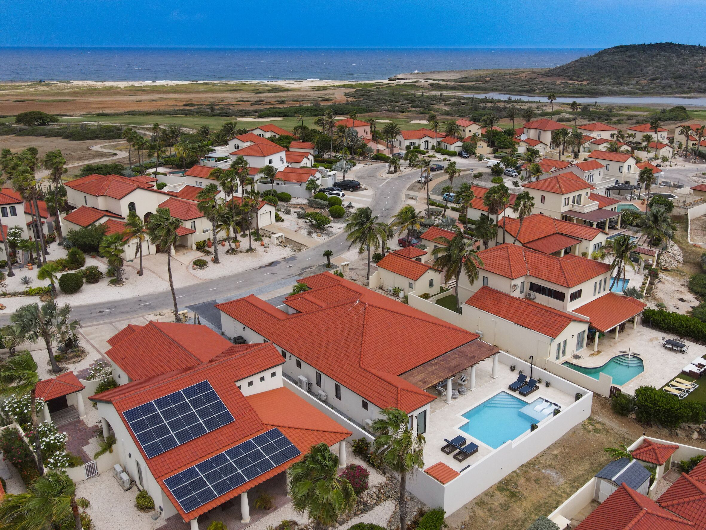A coastal residential area with houses featuring red-tiled roofs, a swimming pool, and solar panels.