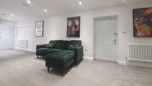 Une pièce spacieuse avec un canapé vert, des murs blancs et des luminaires au plafond. Des tableaux encadrés ornent les murs, et il y a un radiateur blanc.