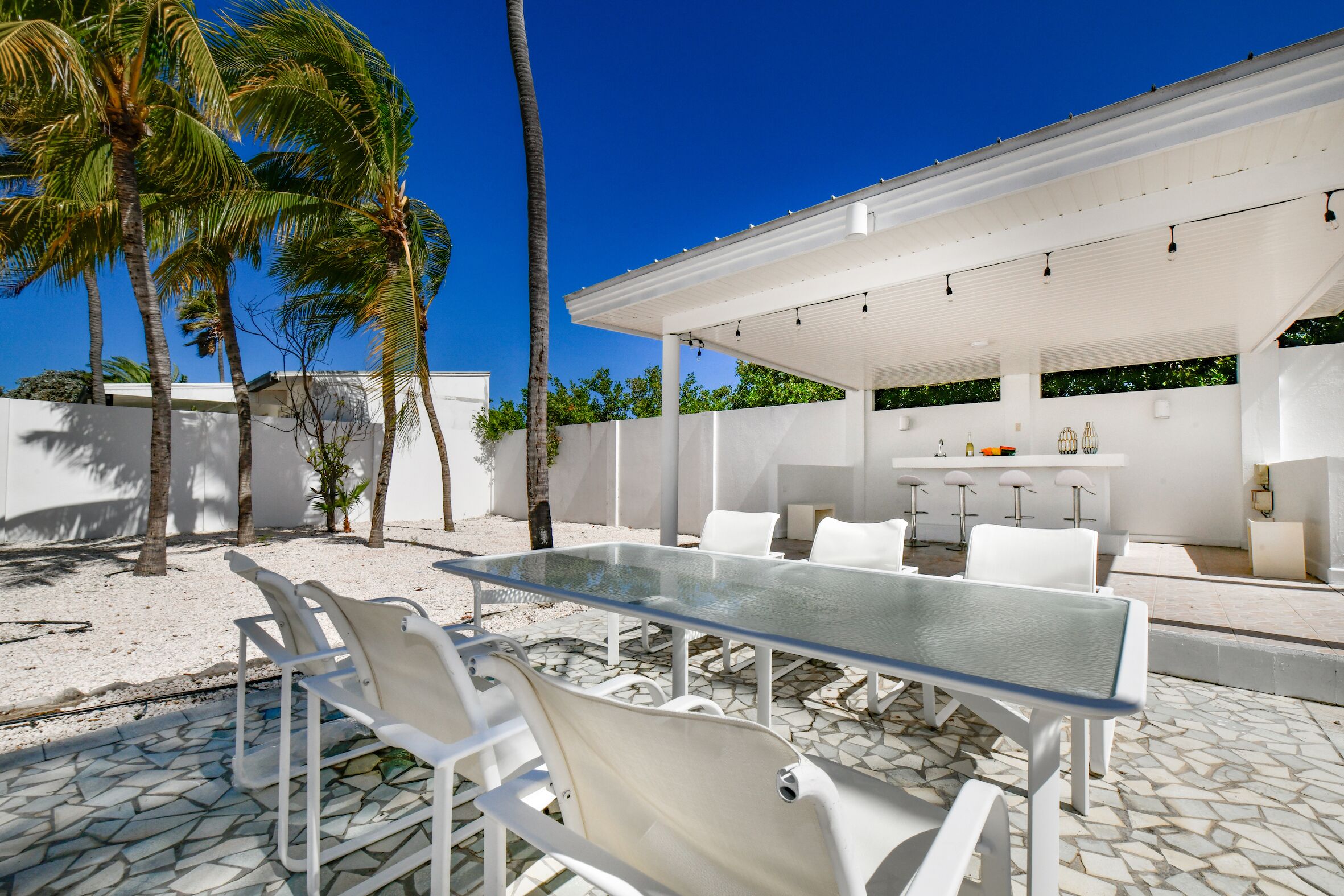 An outdoor patio with a glass-top table and white chairs, surrounded by palm trees and a white building with a bar area.