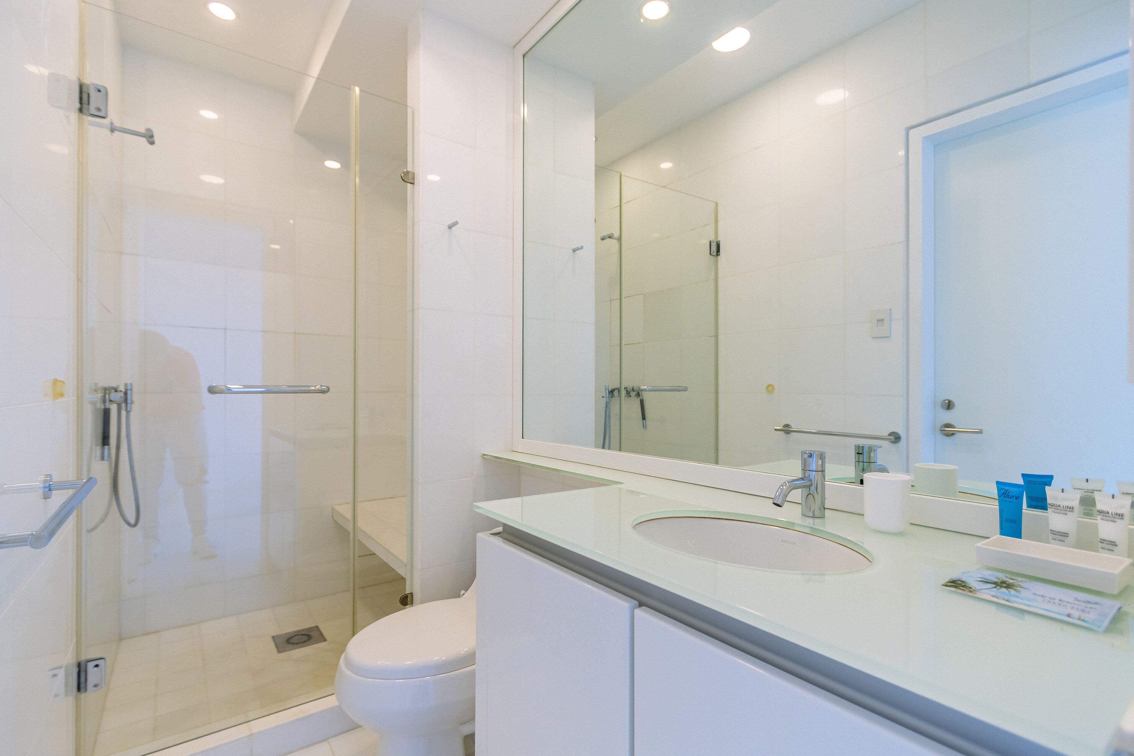 A modern bathroom with a glass shower enclosure, a white toilet, and a large mirror above a sink with toiletries.