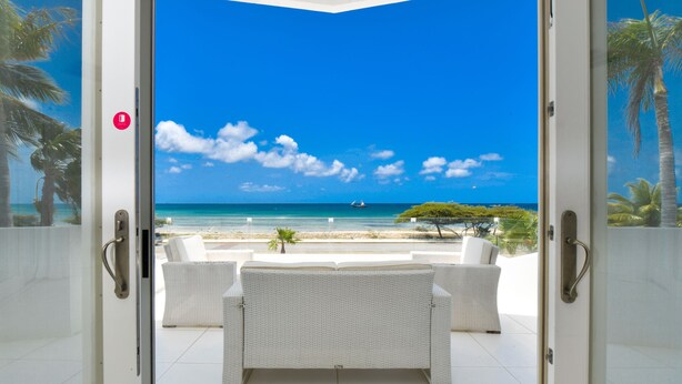 Un balcón con mobiliario de mimbre blanco, vista despejada a la playa y una palmera.