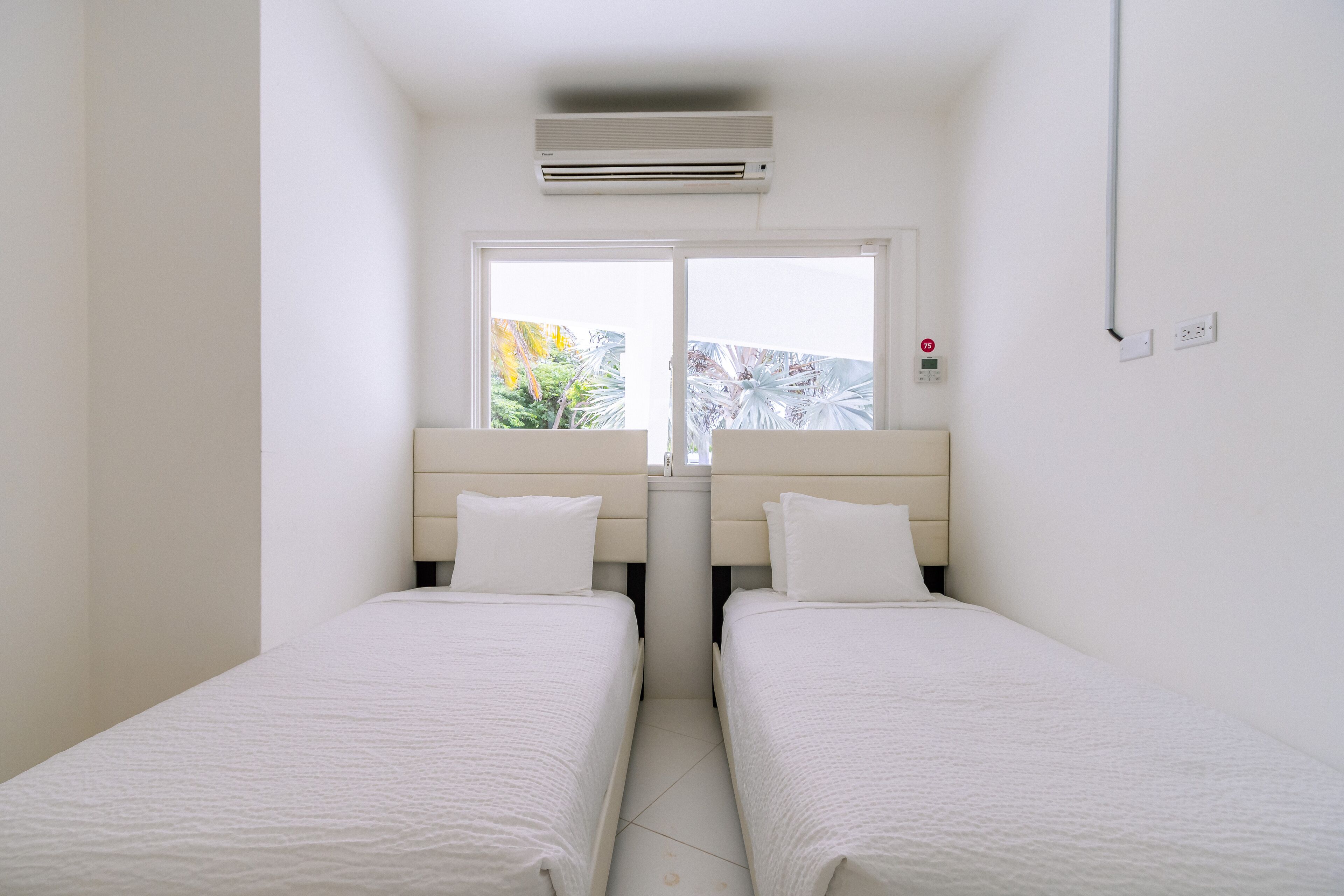 A small, clean room with two single beds, a window with a view of greenery, and an air conditioning unit above the beds.