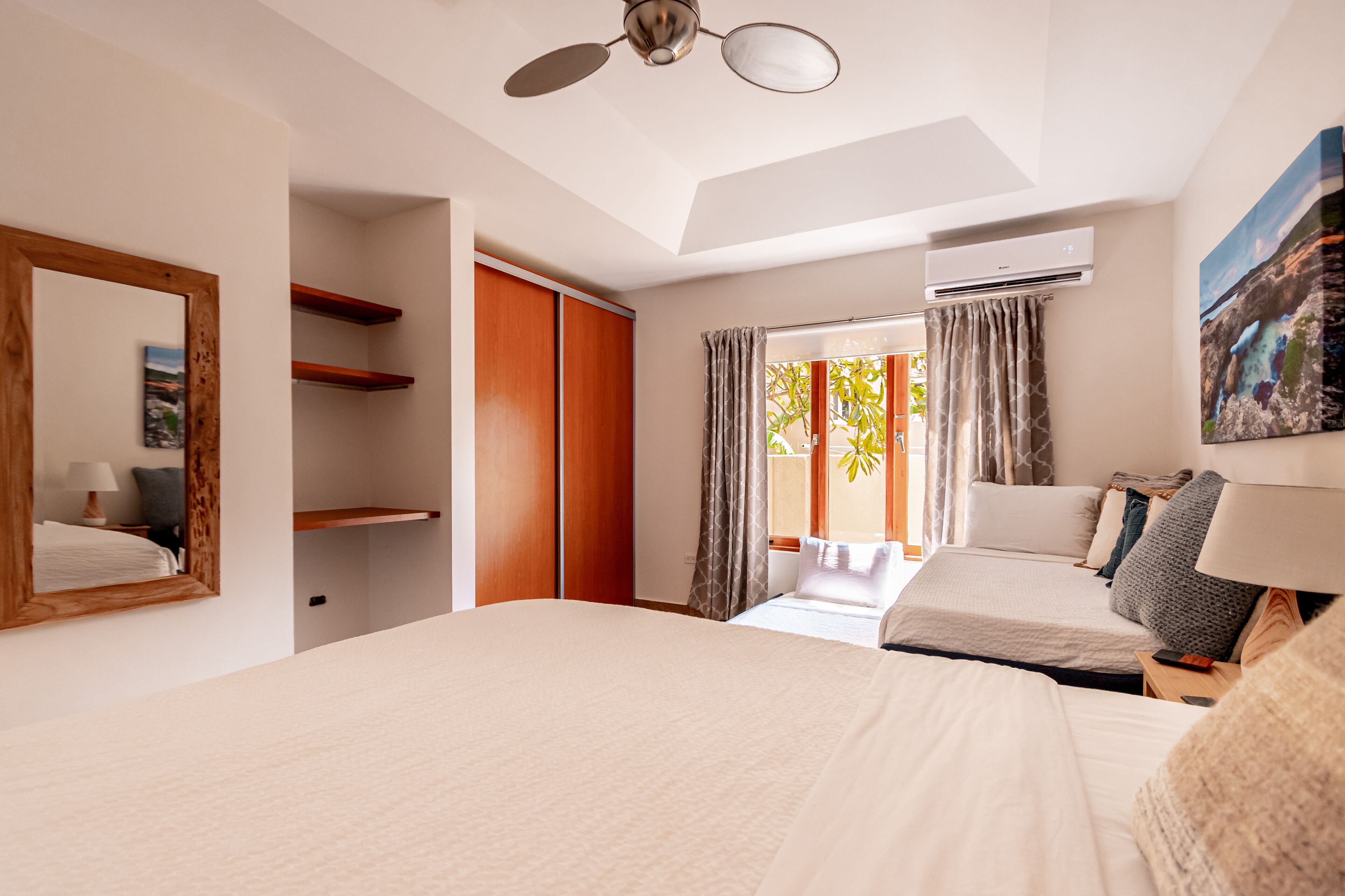 A hotel room with a large bed, a wardrobe, a mirror, a ceiling fan, and a window with curtains.