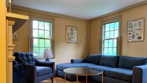 A living room with a blue sofa, a matching armchair, and a glass coffee table. There are two windows with shutters and wall art depicting animals. - Maple Suite- Private  Ground Floor Apartment in front of Smith College (Northampton)