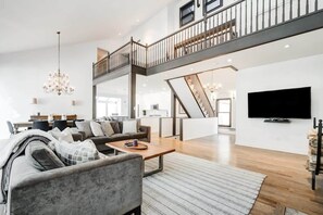 A spacious living area with a sofa set, a coffee table, and a chandelier. A TV is mounted on the wall, and there is a staircase leading to an upper level. - Waterfront open concept dream cottage with hot tub (Portland)
