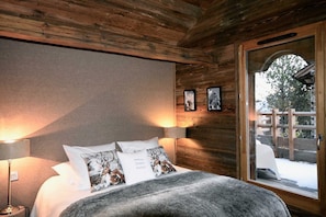Une chambre confortable avec un plafond en bois, un grand lit recouvert de draps blancs, deux lampes fixées au mur et une fenêtre donnant sur un paysage enneigé.