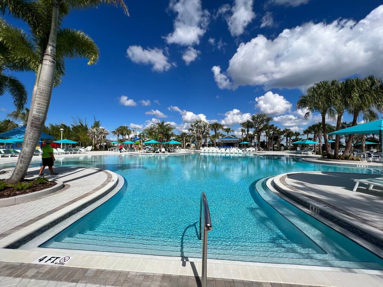 A large, clear swimming pool with a 4 FT depth marker, surrounded by palm trees and lounge chairs under blue umbrellas.