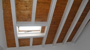 Ein Zimmer mit schrägem Dach, Holzbalken und einem Fenster.