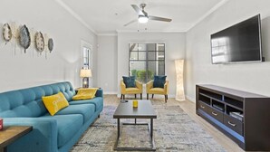 Una sala de estar con un sofá azul, dos sillones amarillos, una mesita de centro, un televisor y un ventilador de techo.