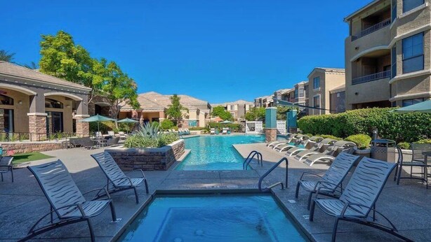 Un área de piscina con sillones de descanso, una piscina azul cristalina y edificios circundantes.