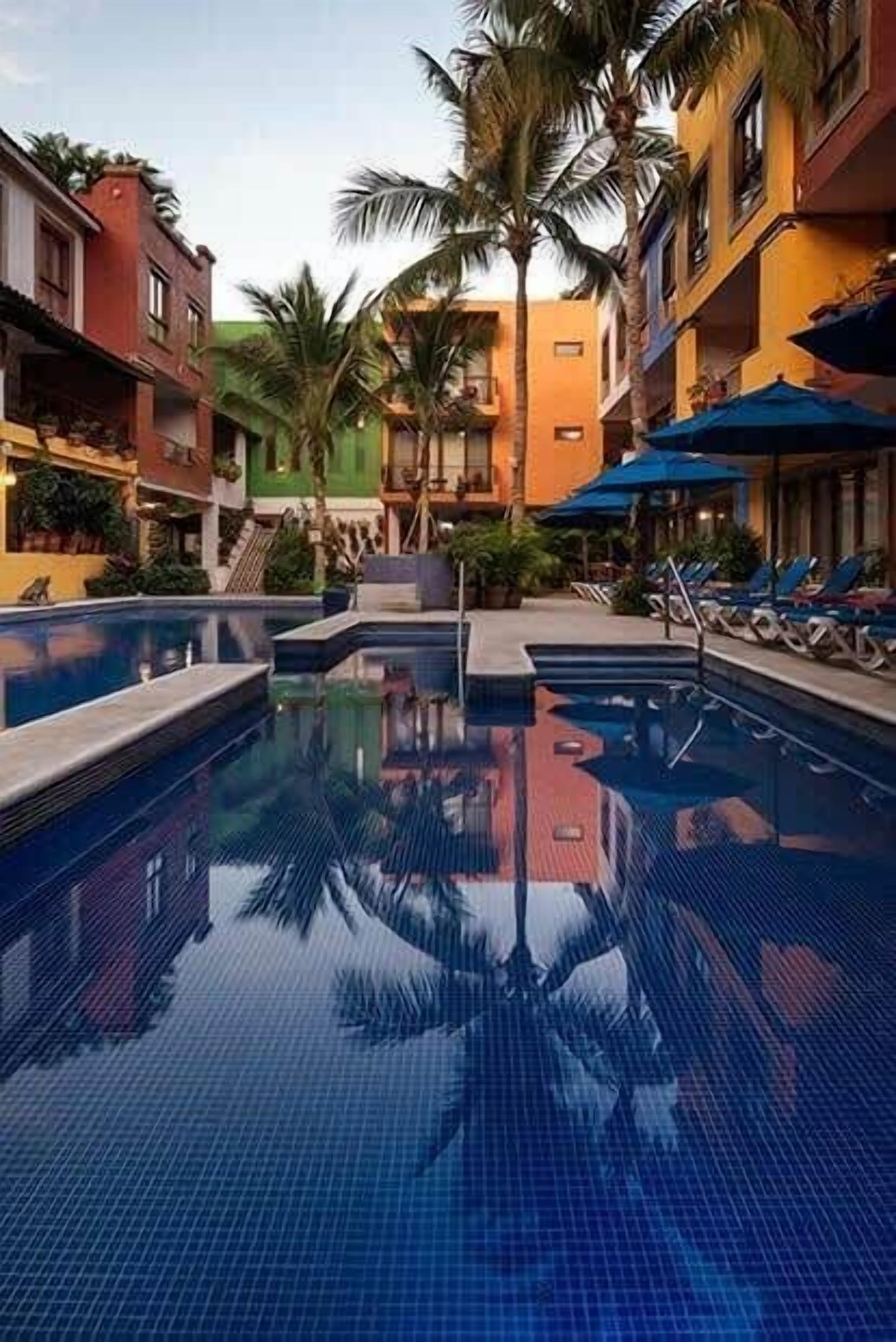 A swimming pool with lounge chairs and umbrellas, surrounded by multi-story buildings with balconies.