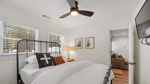 A bedroom with a bed, a ceiling fan, a window with blinds, a bedside table with a lamp, and a door leading to another room.