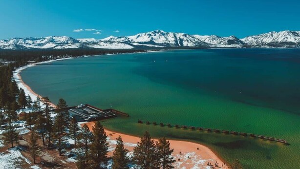 A coastal area with a pier extending into turquoise waters, surrounded by snow-capped mountains and a small beach area.