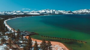 A coastal area with a pier extending into turquoise waters, surrounded by snow-capped mountains and a small beach area.