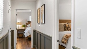 A hallway leading to a bedroom with a bed, a nightstand, and a lamp. There are framed pictures on the wall and a window with blinds.