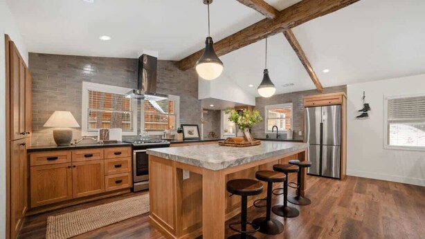 A spacious kitchen with wooden cabinets, a large island with stools, a stainless steel refrigerator, and a farmhouse-style ceiling with exposed beams.