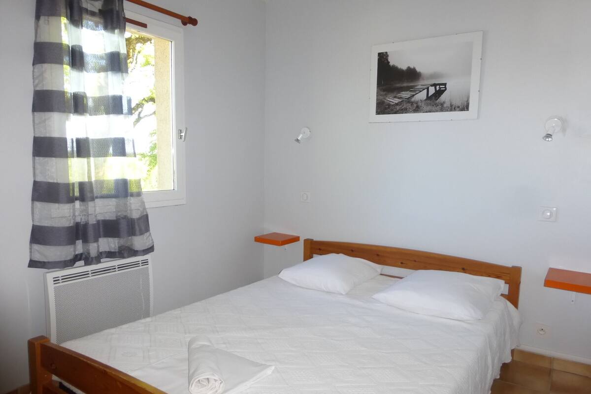 Un dormitorio con cama de madera, ropa de cama blanca, una pequeña estantería, un radiador y un cuadro en la pared.