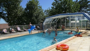 Une piscine avec des personnes dans l’eau, un espace de repos couvert et une terrasse clôturée avec des chaises longues.