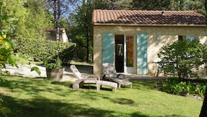 Une petite maison avec un toit en tuiles, une porte et une fenêtre. Dans la cour, on trouve deux fauteuils et un pot de fleurs.