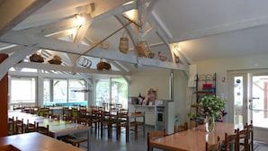 Un espace repas avec des tables et des chaises en bois, un coin cuisine équipé d’un évier et de placards, et un plafond avec des poutres apparentes et des paniers suspendus.