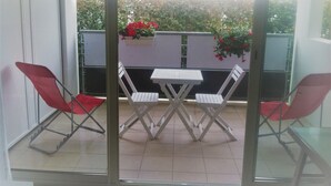 Een balkon met een glazen deur, twee witte klapstoelen, een klein tafeltje en rode stoelen.