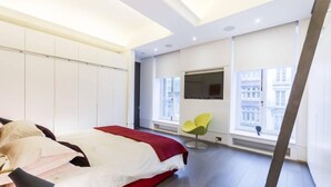 Ein modernes Schlafzimmer mit einem großen Bett, einem Fernseher, einem Fenster mit Blick auf Gebäude und einem leuchtend gelben Sessel.