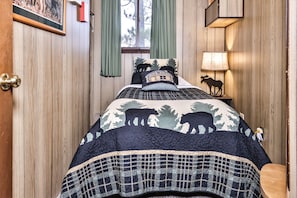 A bedroom with a bed featuring a quilt with animal prints, a wooden door, a window with curtains, and a bedside lamp.