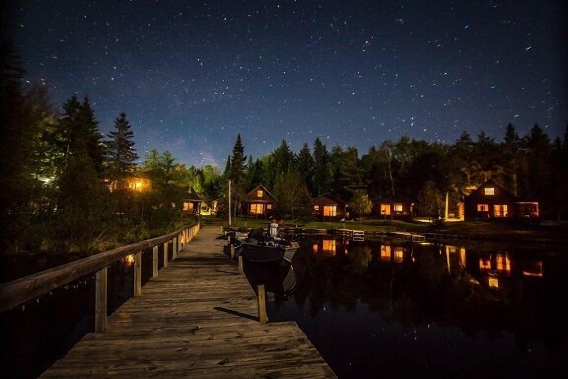 A wooden dock extending into a calm body of water with houses on the opposite shore, under a starry night sky.