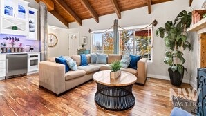 A modern living room with a wooden ceiling, a beige sectional sofa, a round coffee table, and a large potted plant. - Luxury Home—Hot Tub, Fire Table, Office, Stunning Lake Views, 15 Miles to Silverwood, 2 to Beach! (Hayden)