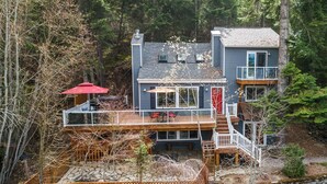 A two-story house with a deck, surrounded by trees. - Luxury Home—Hot Tub, Fire Table, Office, Stunning Lake Views, 15 Miles to Silverwood, 2 to Beach! (Hayden)