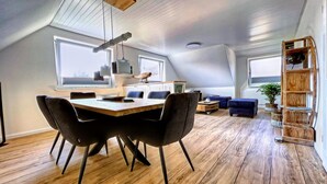Un loft espacioso con techo alto, un comedor con mesa y sillas de madera, una sala de estar con sofá, y una estantería.