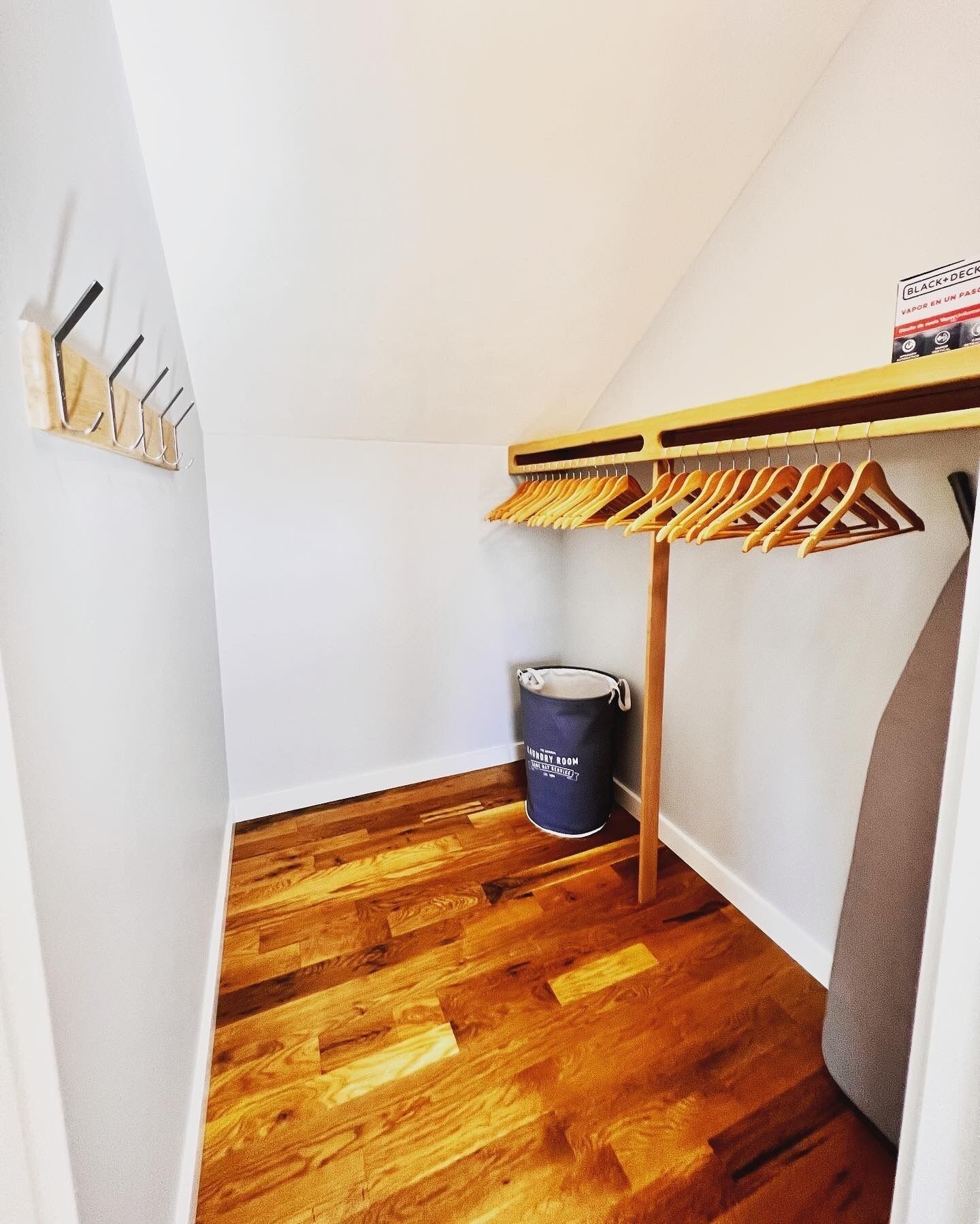 A small, narrow room with wooden flooring, a wooden coat rack, a blue bucket labeled "Laundry Room," and a white wall.