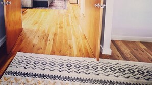 Una habitación con piso de madera, una cama con cabecera gris, una mesita con una planta y un ventanal con vista a follaje.