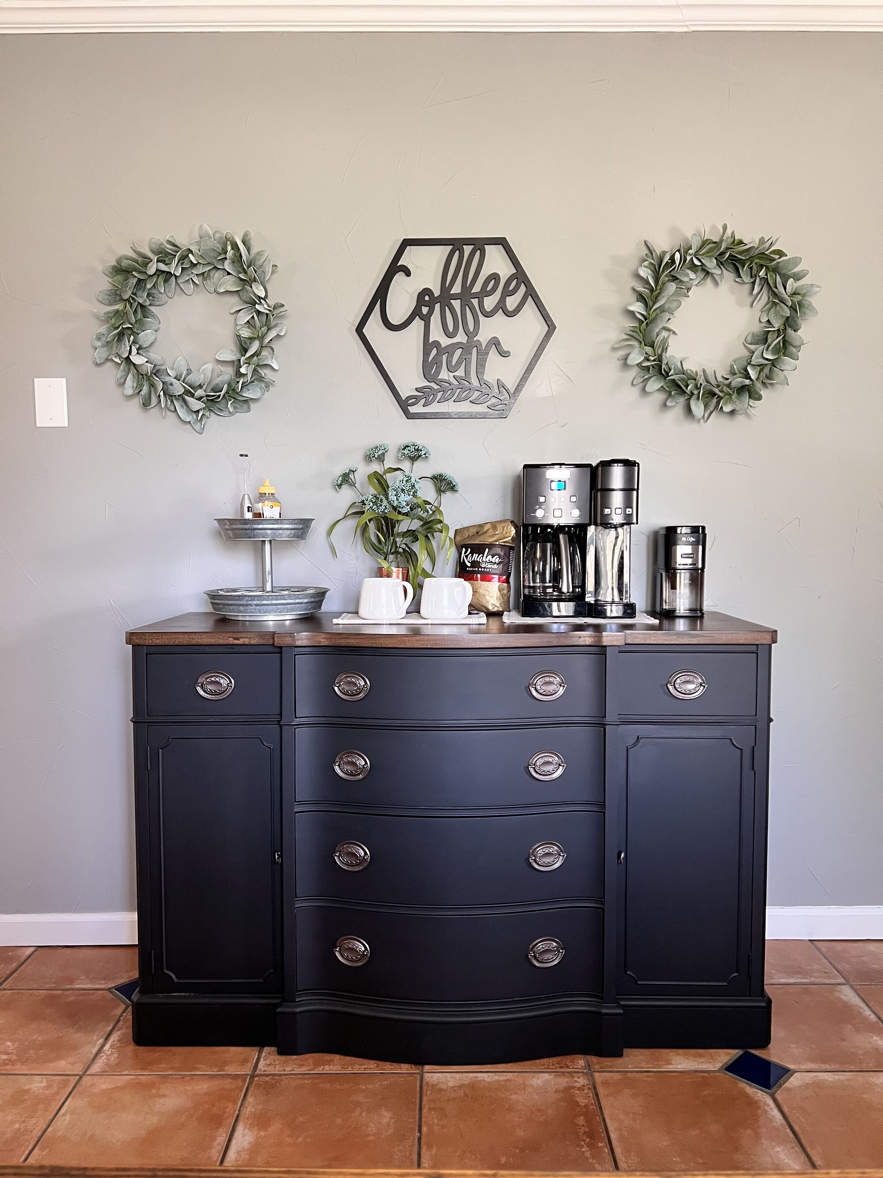 A kitchen counter with a coffee maker, a mug, and a bag of coffee. There are two wall decorations, one saying "Coffee" and the other "Coffee bar".