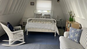 Um quarto com teto inclinado, uma cama, uma cadeira de balanço, uma mesinha de cabeceira, uma estante de livros e uma janela com persianas.