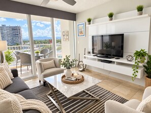 Un salon moderne avec un canapé, une table basse et un téléviseur. Il y a un balcon avec des chaises et une vue sur un immeuble.