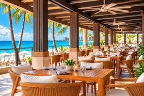A covered outdoor dining area with tables and chairs, palm trees, and a view of the ocean.