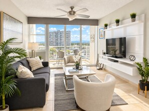 Un salon moderne avec un canapé, un fauteuil, une table basse et un téléviseur. Il y a un balcon avec vue sur les immeubles et un ventilateur de plafond.