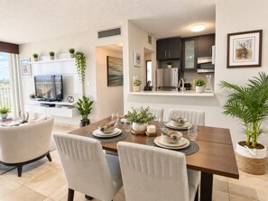 Una moderna zona pranzo con un tavolo in legno apparecchiato per quattro persone, circondato da sedie. Sullo sfondo è visibile una cucina con mobili scuri e elettrodomestici in acciaio inossidabile.
