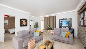Ein modernes Wohnzimmer mit zwei grauen Sofas, einem Couchtisch und einem Fernseher. Im Hintergrund befindet sich ein Bücherregal und eine Pflanze.