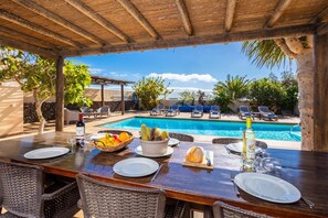 Comedor con vista a la piscina, asientos al aire libre y una mesa para dos, con frutero y botella de vino.