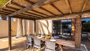 Eine Terrasse im Freien mit Holztisch und -stühlen, einem Reetdach und einem Pool im Hintergrund.