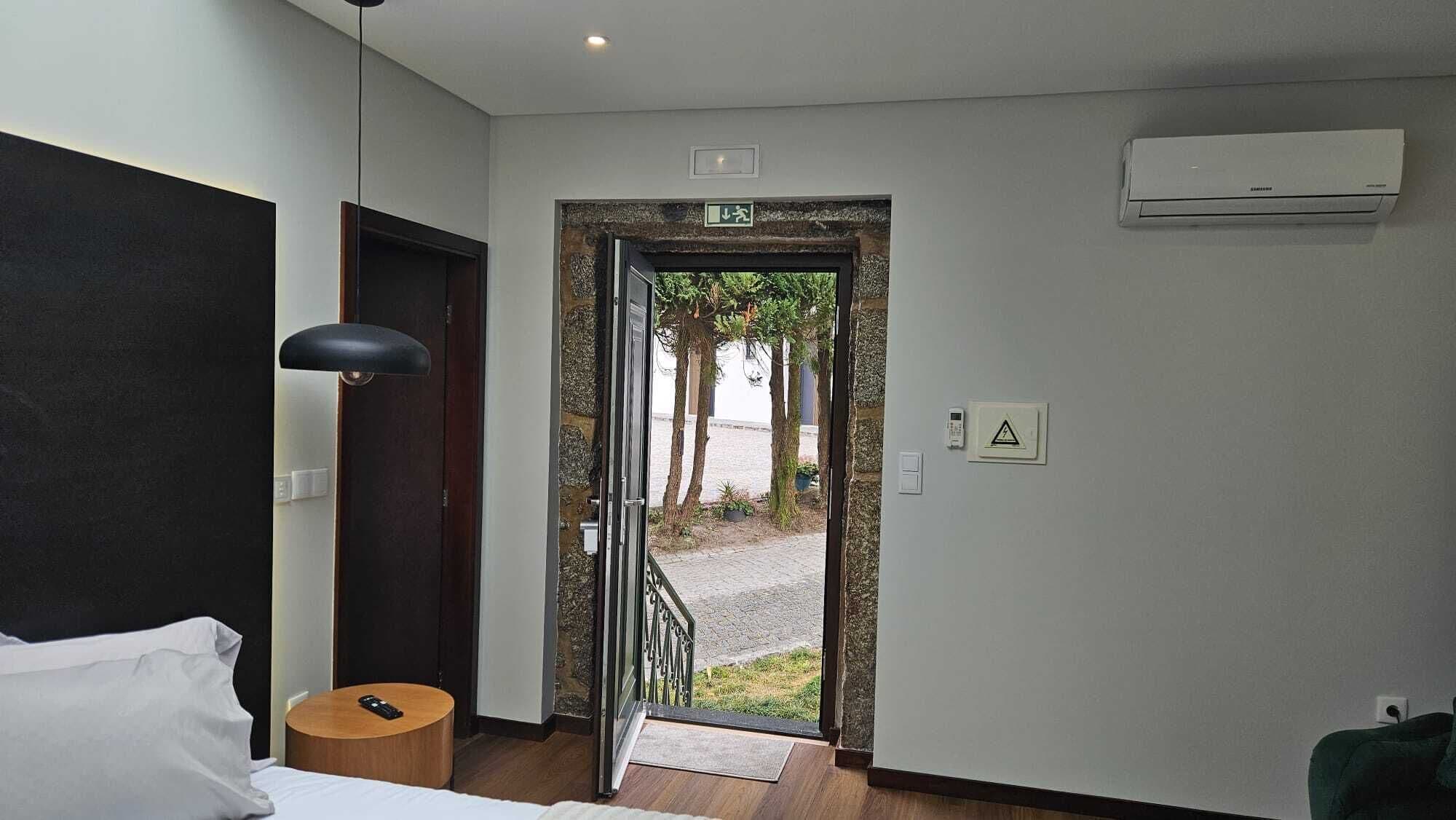 A hotel room with a bed, a desk, a chair, a door leading to an outdoor area with trees, and an air conditioning unit on the wall.
