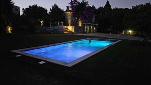 A rectangular swimming pool with a person swimming, surrounded by a well-lit garden and a stone building with steps leading up to it. - ICH Inveja Country House (Pacos De Ferreira)